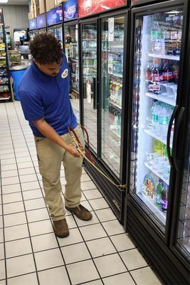 A person in a blue polo shirt and khaki pants holds a measuring tape while working in front of a row of store refrigerators.
