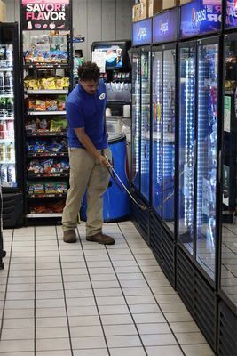 A person in a blue shirt and tan pants uses a long pole to clean the glass doors of a refrigerated aisle in a store.