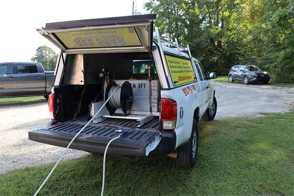 A white pickup truck with an open camper shell containing a hose reel and equipment, parked on a grassy area.