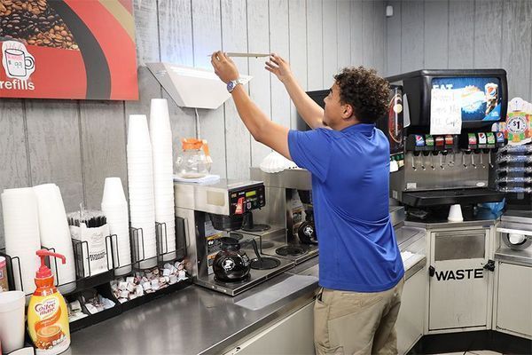 A person in a blue shirt reaches up to adjust an object attached to a light-colored wall in a beverage station.