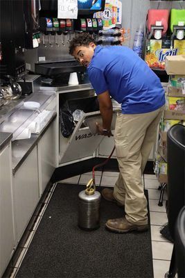 A worker in a blue shirt and tan pants uses a metal pest control sprayer behind a soda fountain counter.