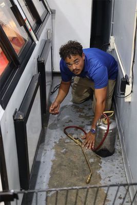 A person in a blue shirt kneels in a narrow space, using a handheld metal spray canister to treat the floor.