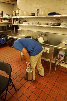 A person in a blue shirt and khaki pants stands in a commercial kitchen, holding a metal pesticide sprayer.