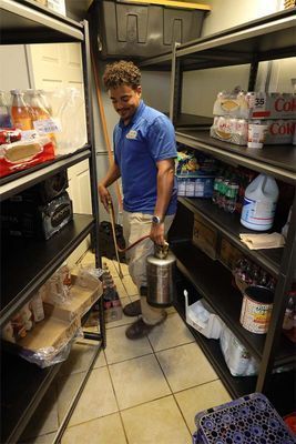 A service professional wearing a blue shirt stands in a storage room, holding a handheld silver pesticide sprayer.