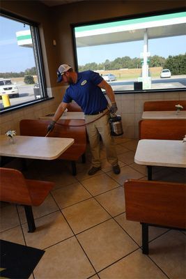 A person wearing a blue shirt and cap sprays disinfectant on booth seats inside a restaurant.