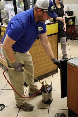 A pest control technician in a blue shirt sprays along the base of a commercial counter using a handheld tank sprayer.