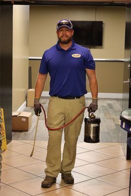 A pest control technician in a blue polo and khaki pants stands indoors holding a pressurized chemical sprayer.