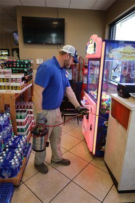 A person in a blue shirt and cap sprays insecticide around a claw machine in a retail store.