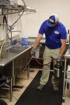 A person in a blue uniform sprays a solution under a commercial kitchen sink using a handheld pressurized metal tank.