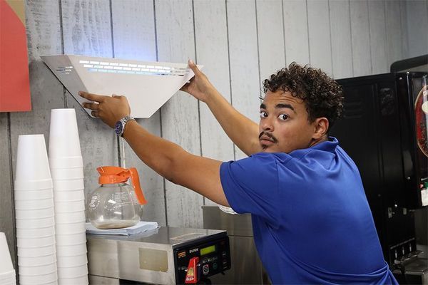 A person in a blue shirt installing or adjusting a white light fixture on a wall above a coffee machine.