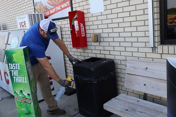 A person in a blue shirt and cap sprays cleaner onto a black outdoor trash can near a brick wall and wooden bench.