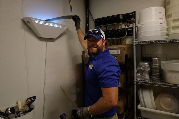 A smiling technician in a blue polo shirt and safety gloves services a wall-mounted light-based insect trap in a kitchen.