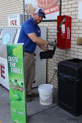 A person in a blue shirt performs maintenance on a fire extinguisher cabinet on a brick wall outside a store.