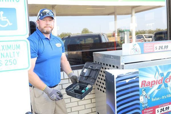A person wearing a blue polo shirt and gloves holds an open tool kit outside next to a propane exchange station.
