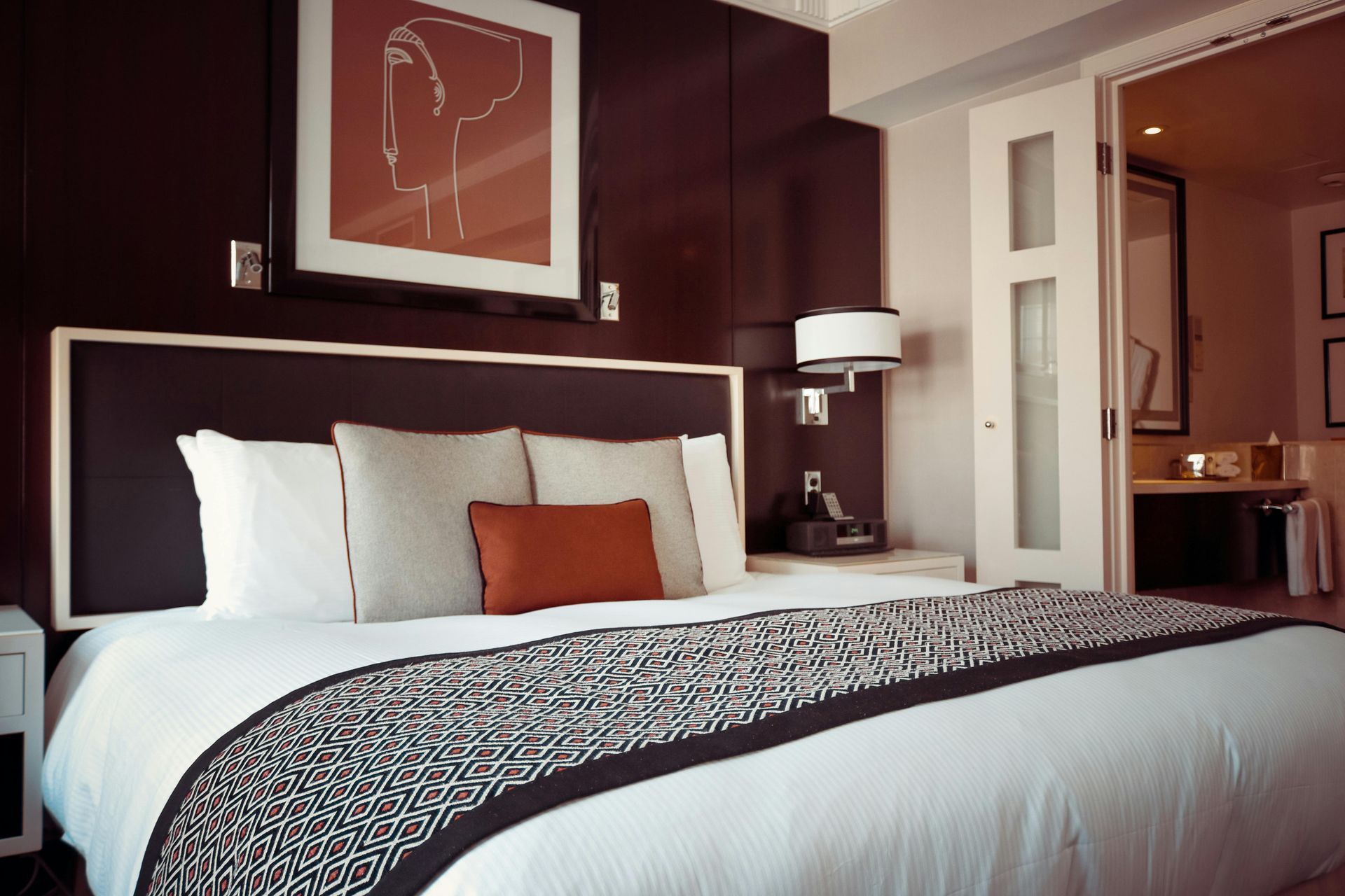 A modern hotel bed with white linens, grey and rust-colored pillows, a patterned runner, and wall art in a dark room.