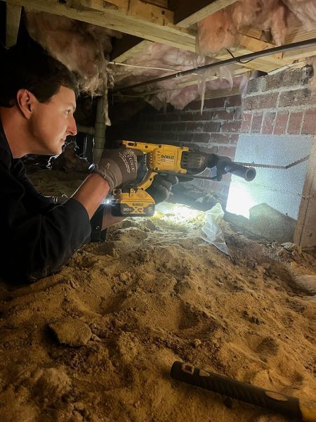 A person in a crawlspace uses a yellow power drill on a brick wall, with exposed insulation visible on the ceiling.