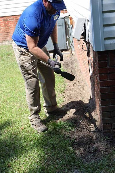 A technician in a blue polo shirt and tan pants uses a tool to dig near the brick foundation of a house.