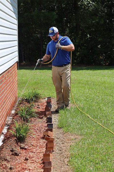 A professional uses a hose to spray treatment along the brick foundation of a house next to a mulch flower bed.