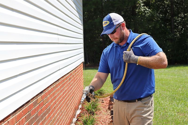 A professional wearing a blue polo and hat sprays a chemical treatment along the brick foundation of a house.