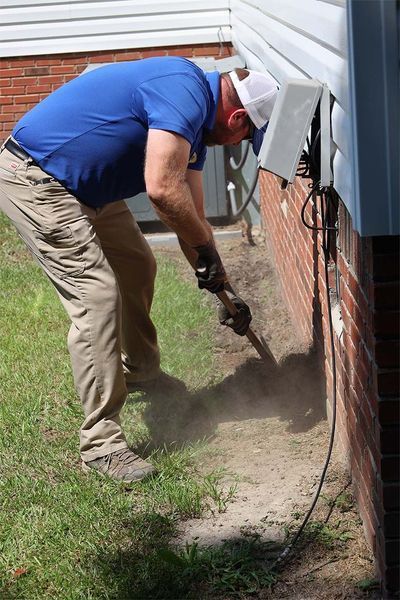 A worker in a blue shirt digs in the dirt near the exterior brick wall of a house where an electrical box is mounted.
