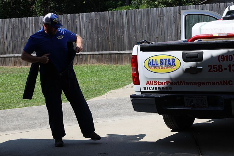 A pest control technician in a blue shirt walks near an All Star Pest Management truck parked on a driveway.