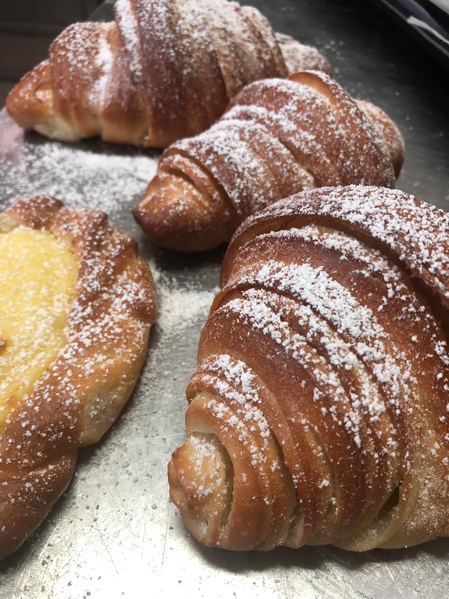 Quattro croissant, due spolverati con zucchero a velo e uno farcito con crema pasticcera, su una superficie metallica.