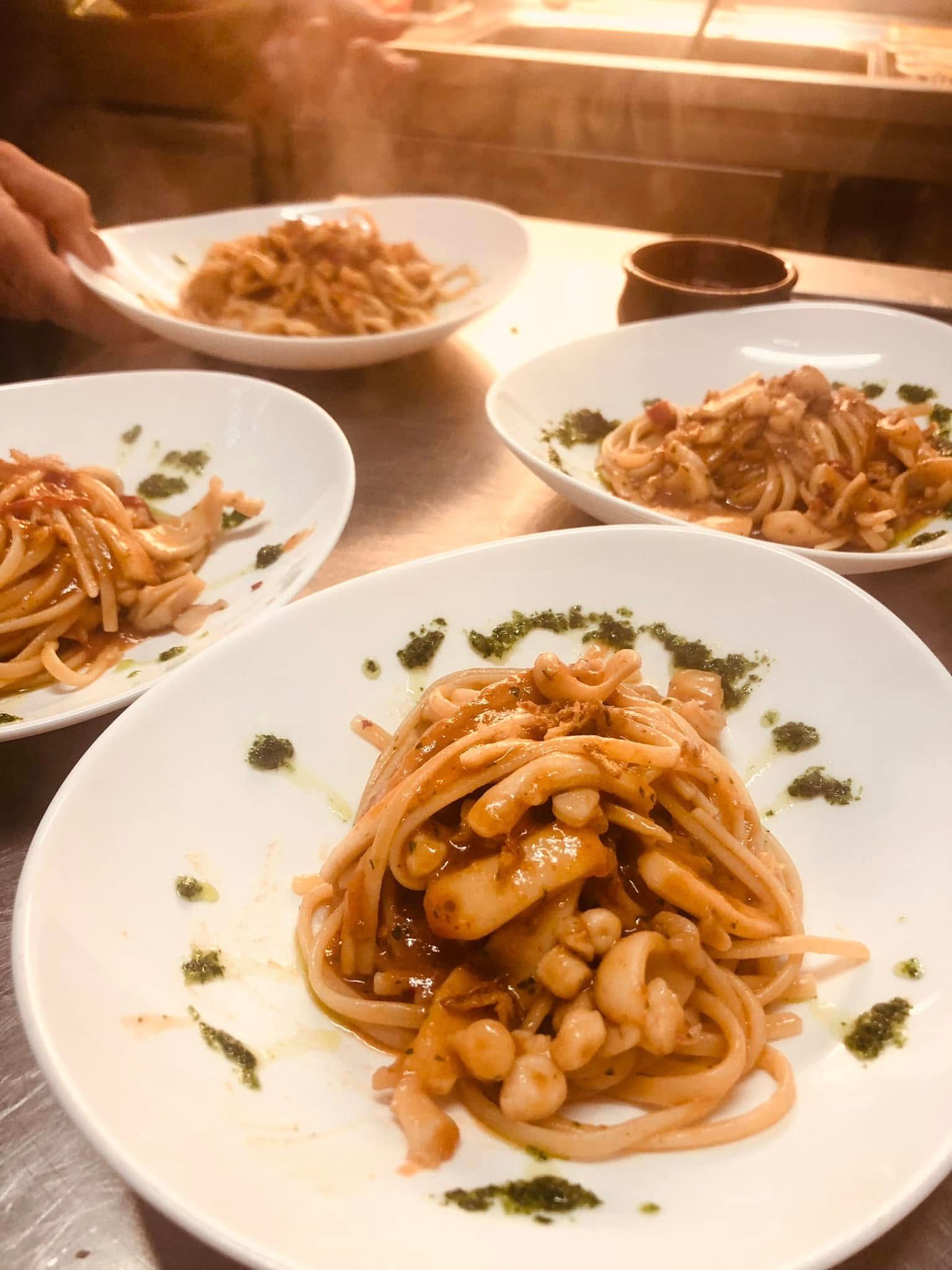 Quattro piatti di pasta conditi con salsa e erbe aromatiche, disposti su una superficie metallica; una mano regge un piatto.