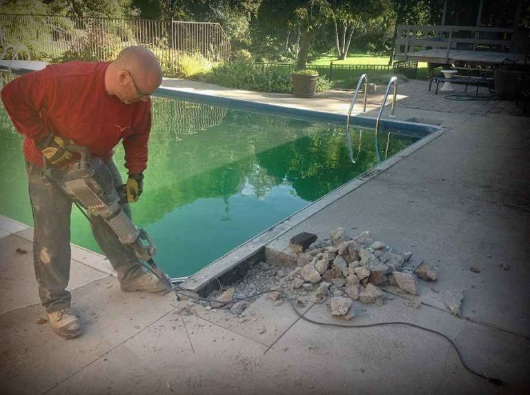 Une personne vêtue d'une chemise rouge à manches longues utilise un marteau-piqueur pour démolir la dalle de béton d'une piscine près d'une piscine verte.