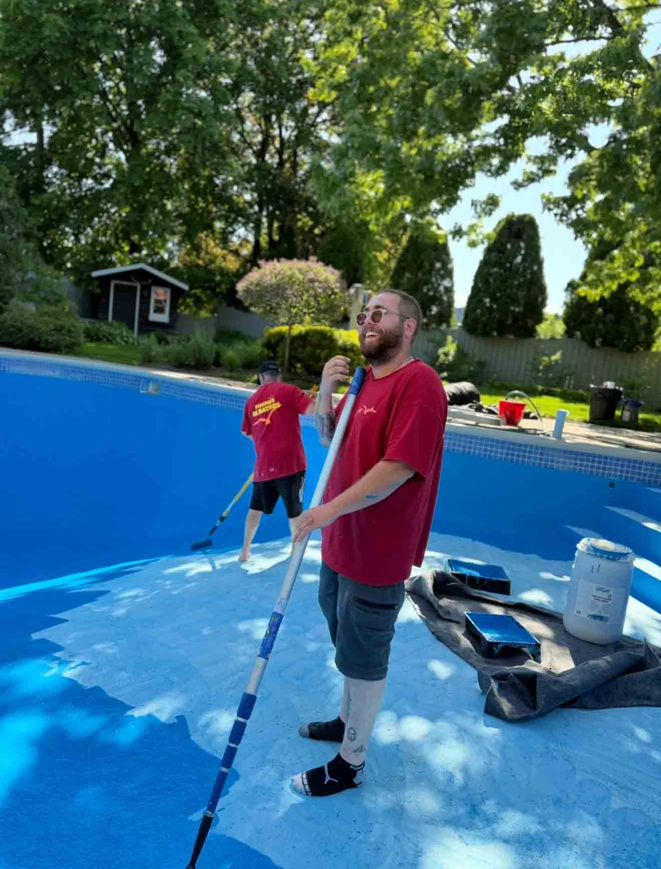 Deux personnes vêtues de chemises rouges utilisent de longues perches pour appliquer une substance blanche sur le sol bleu d'une piscine extérieure vide.