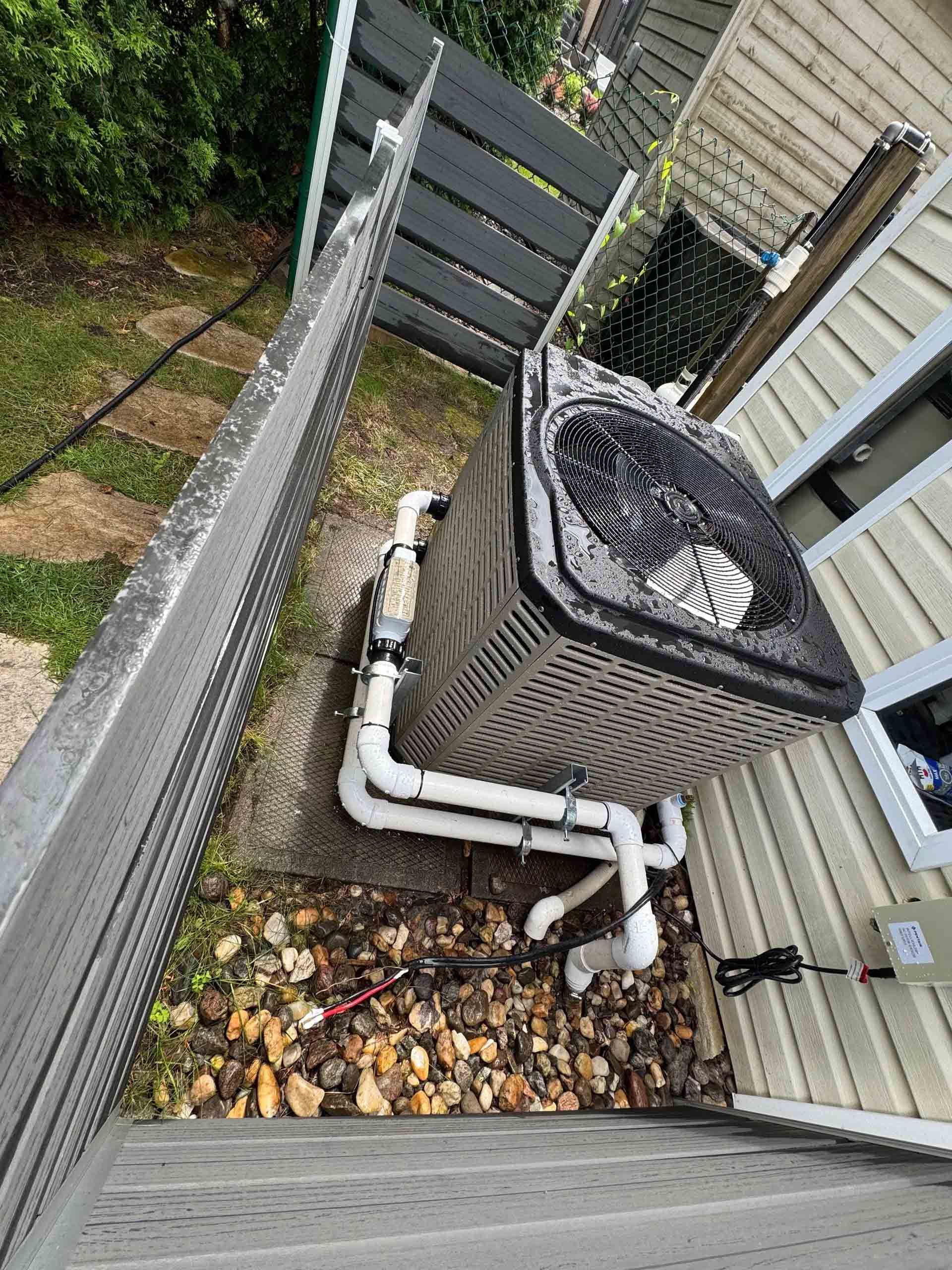 Unité de climatisation extérieure avec tuyauterie en PVC blanc, située sur une surface gravillonnée à côté d'une maison beige et d'une clôture sombre.