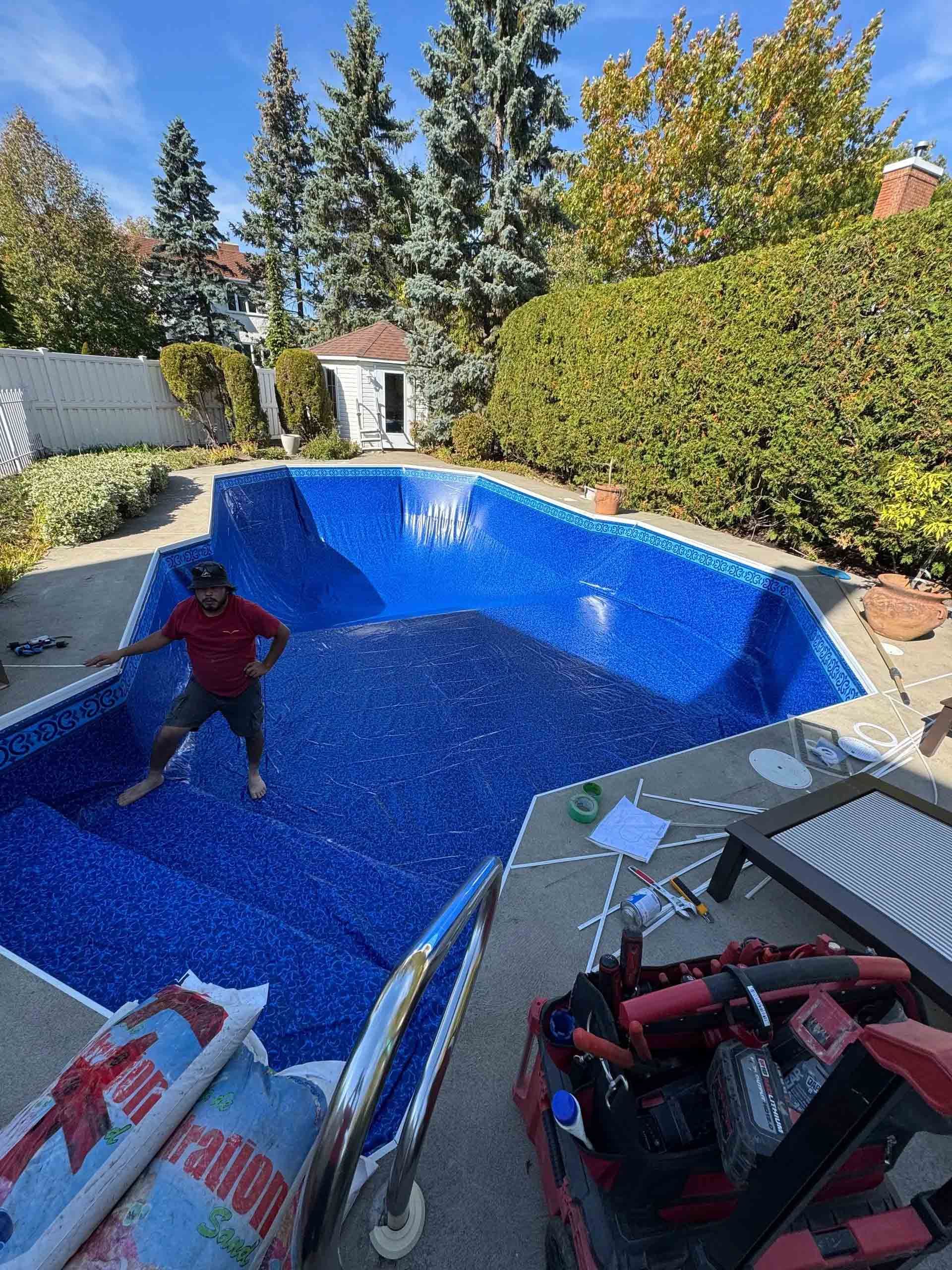 Un ouvrier se tient debout dans une piscine vide, carrelée de bleu, récemment installée dans un jardin ensoleillé bordé de haies vertes.