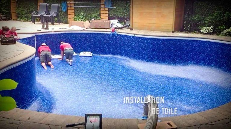 Deux ouvriers installent un revêtement en vinyle bleu dans une piscine vide située dans un jardin.