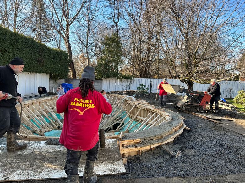 Des ouvriers construisent un rebord de piscine incurvé en béton dans une cour arrière, en coulant du ciment dans des coffrages en bois sur une surface de gravier.