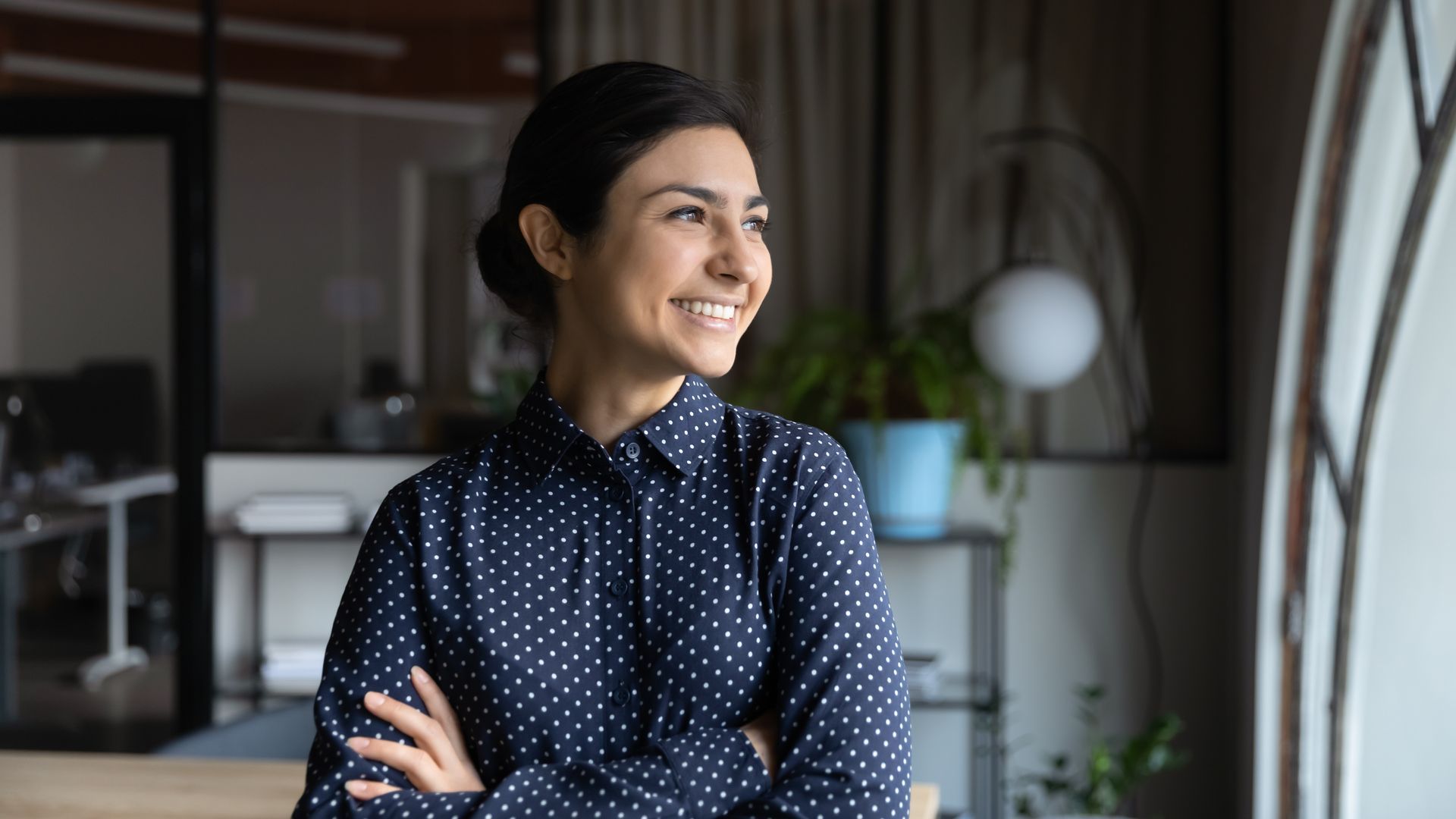 Woman with arms crossed, smiling, looking out a window in an office. She wears a blue, polka-dot shirt.