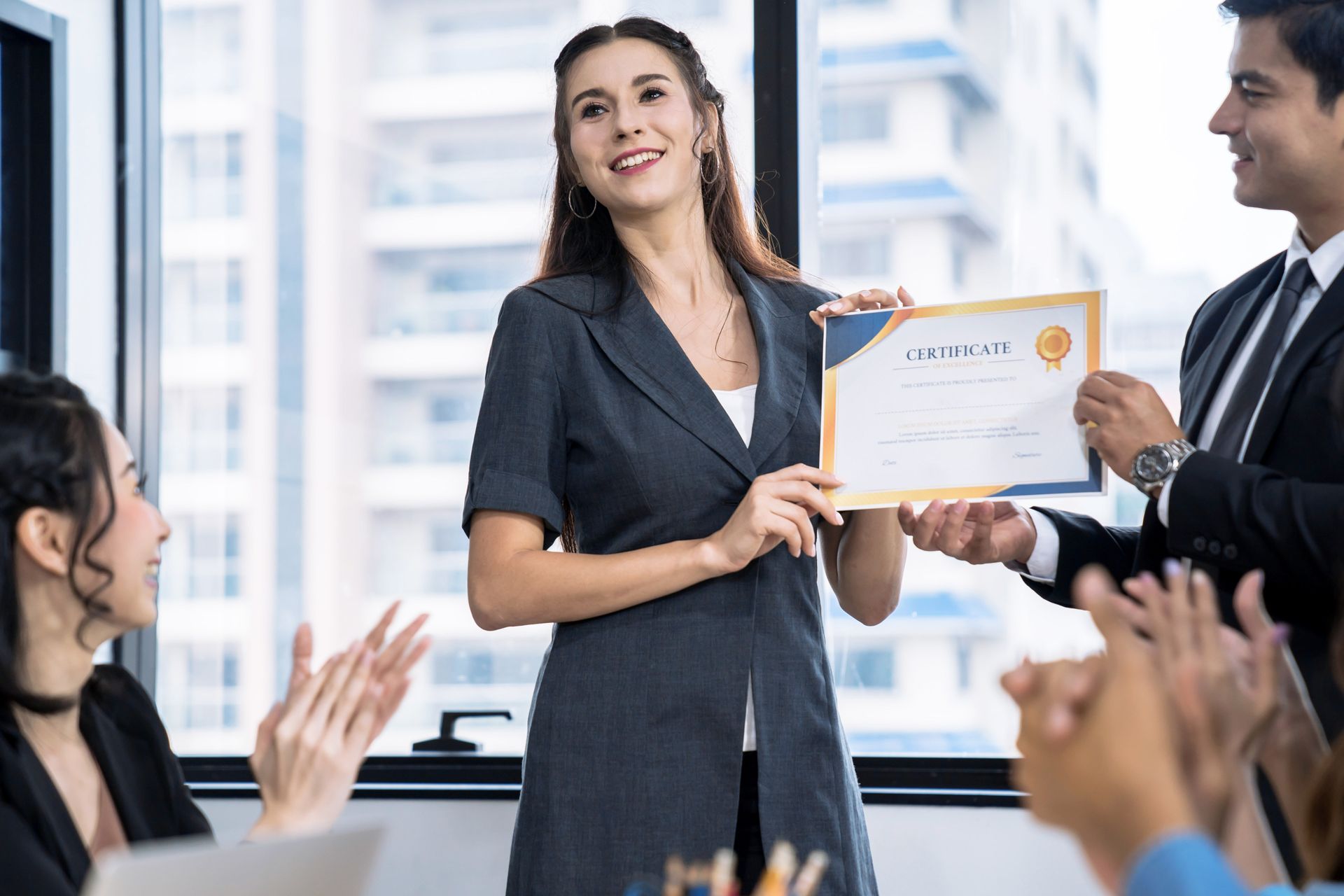 Woman in gray suit receives certificate at work, others applaud.