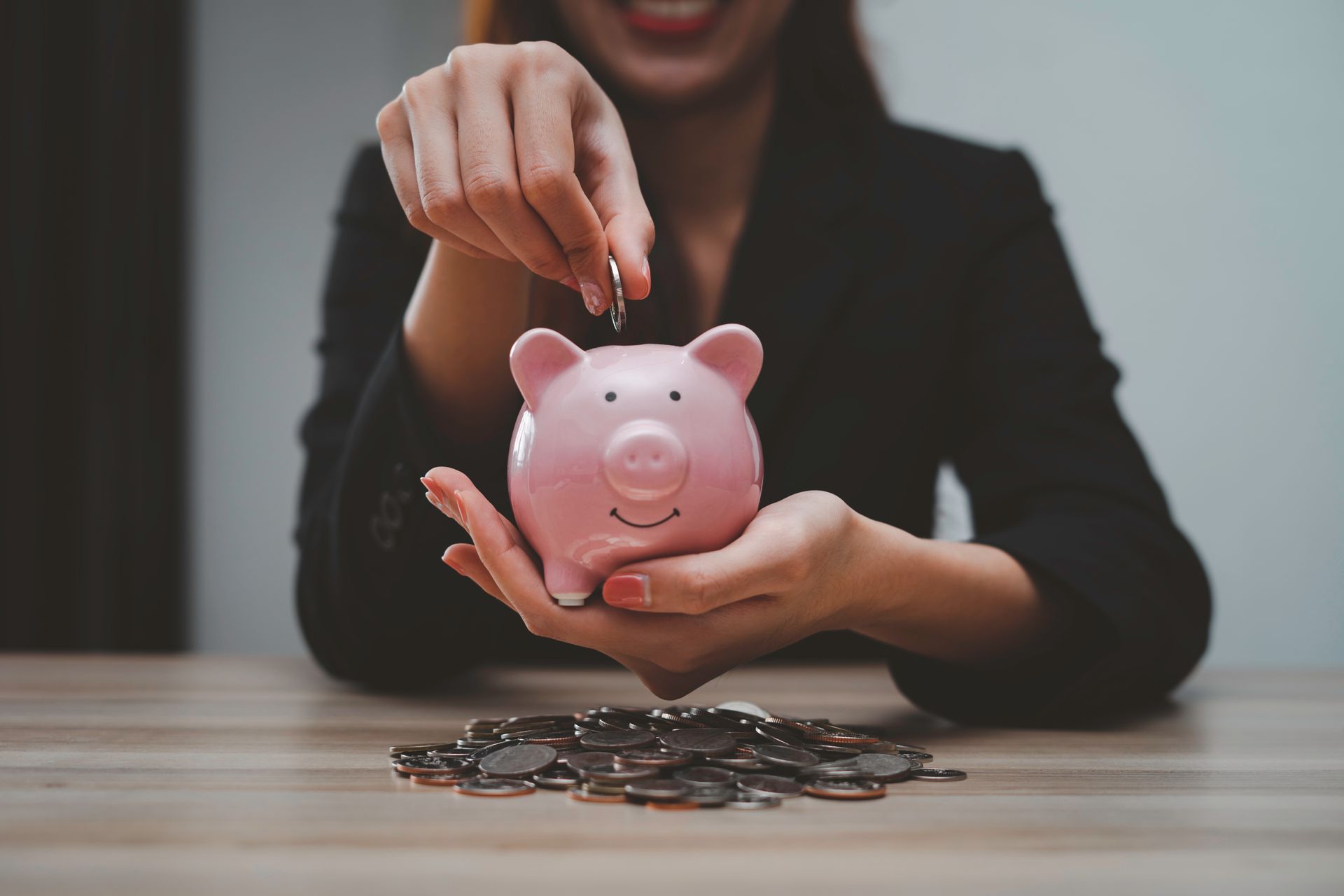 Woman putting a coin into a pink piggy bank on a wooden table with a pile of coins; smiling.
