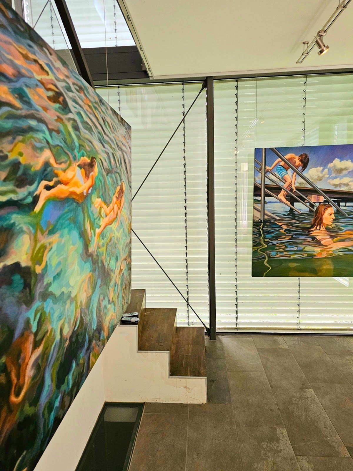 Zwei Gemälde von im Wasser schwimmenden Figuren sind in einer modernen Galerie mit Treppe und großen Fenstern ausgestellt.