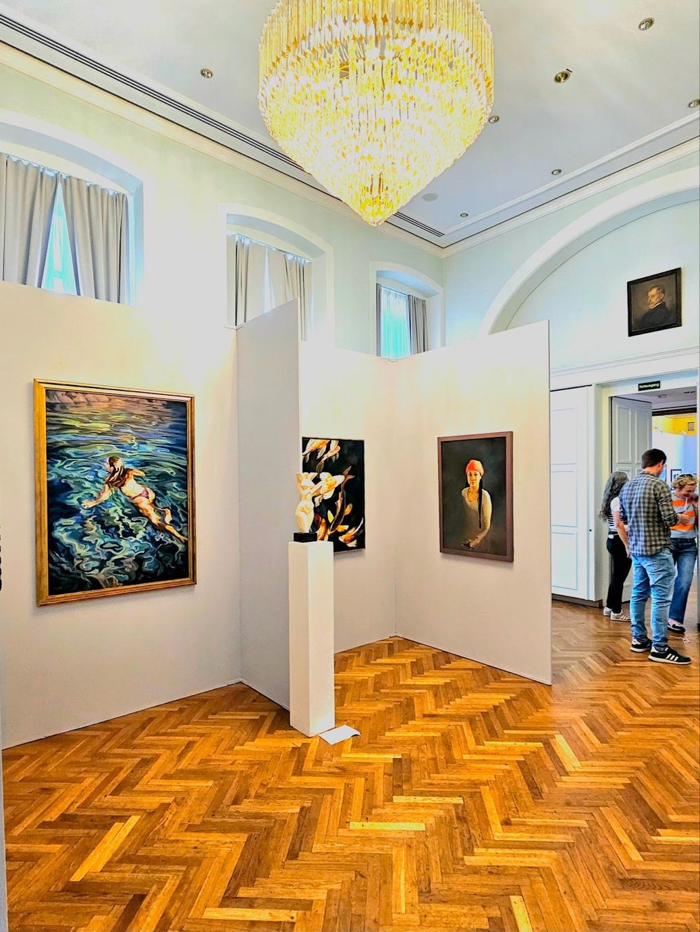 Ein Kunstgalerieraum mit Gemälden an weißen Wänden, Parkettboden im Fischgrätmuster und einem großen Kristalllüster.