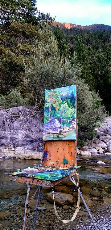 Auf einem felsigen Flussufer vor einem Bergwald steht bei Sonnenuntergang eine Staffelei mit einem Landschaftsgemälde.