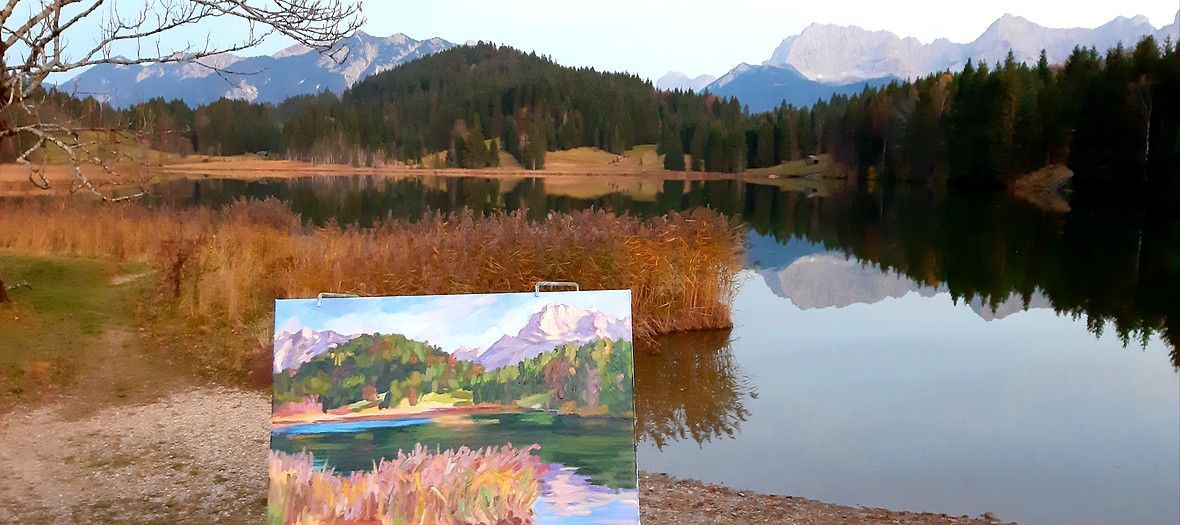 Auf einem Strand steht eine Staffelei mit dem Bild eines malerischen Bergsees.