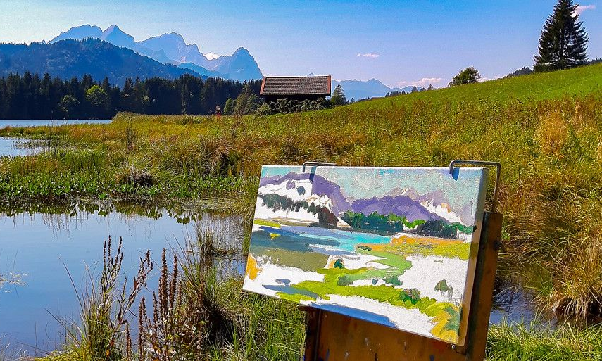 Eine Staffelei mit einem impressionistischen Landschaftsgemälde im Freien, mit Blick auf einen See, grasbewachsene Felder.