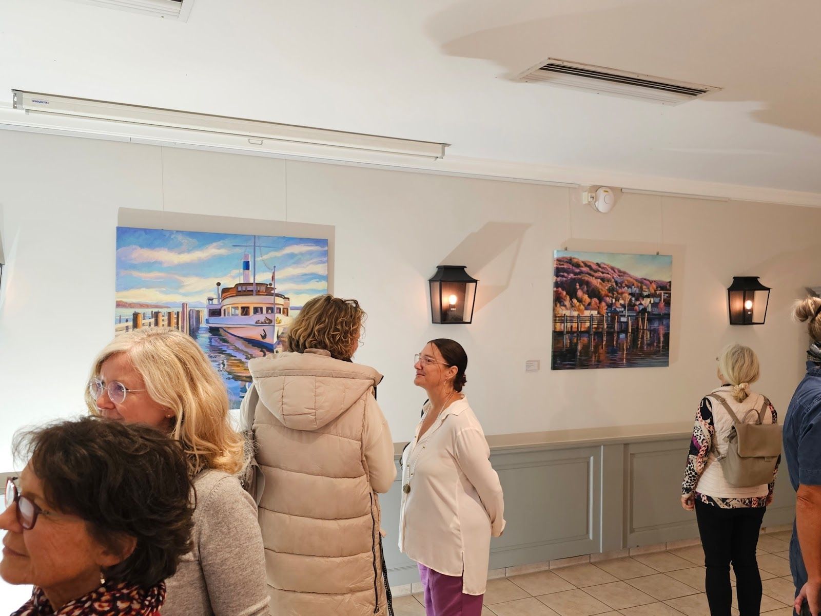 In einer hell erleuchteten Galerie versammelten sich Menschen und betrachteten gerahmte Landschaftsgemälde.