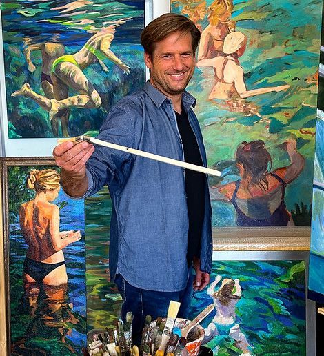 Ein Künstler lächelt, während er einen Pinsel vor mehreren Gemälden hält, die Menschen beim Schwimmen unter Wasser zeigen.