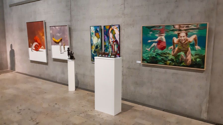 In einer Kunstgalerie sind Gemälde und eine kleine Skulptur auf einer Betonwand und weißen Sockeln ausgestellt.