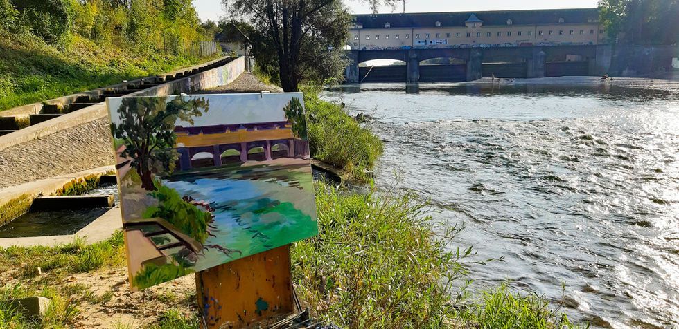 Auf einer Staffelei steht ein Gemälde einer Holzbrücke über einen Fluss, das auf einem grasbewachsenen Ufer.