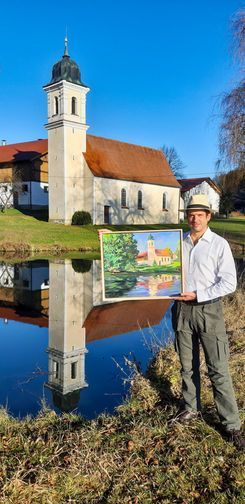 Eine Person hält ein Gemälde einer Kirche an einem Teich in der Hand, das der tatsächlichen Kirche.