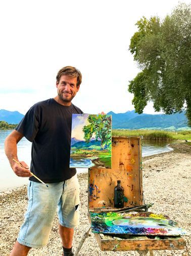 Eine lächelnde Person hält ein Landschaftsgemälde im Freien an einem See in der Hand; Staffelei.