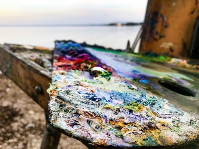 Nahaufnahme einer verwitterten hölzernen Malerpalette, bedeckt mit dicken Schichten bunter Ölfarbe, mit Blick auf einen See.