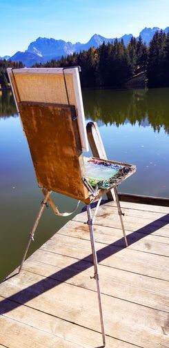 Eine leere französische Holzstaffelei steht auf einem Holzsteg mit Blick auf einen See, im Hintergrund sind Wald.