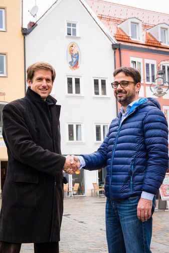 Zwei Männer in Freizeitkleidung schütteln sich im Freien vor einem weißen Gebäude mit Fenstern.